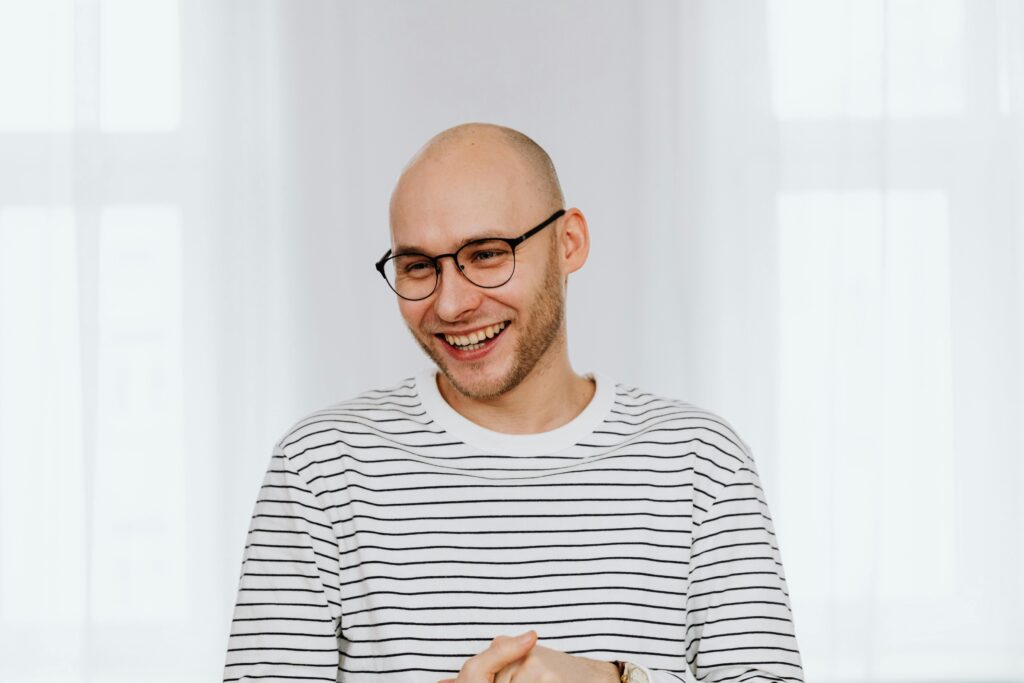 A bald man with glasses and a joyful smile in a striped shirt indoors.